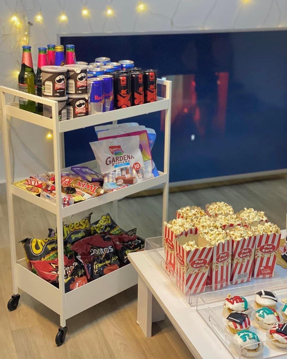 Snack cart and table with popcorn and cupcakes under warm string lights.