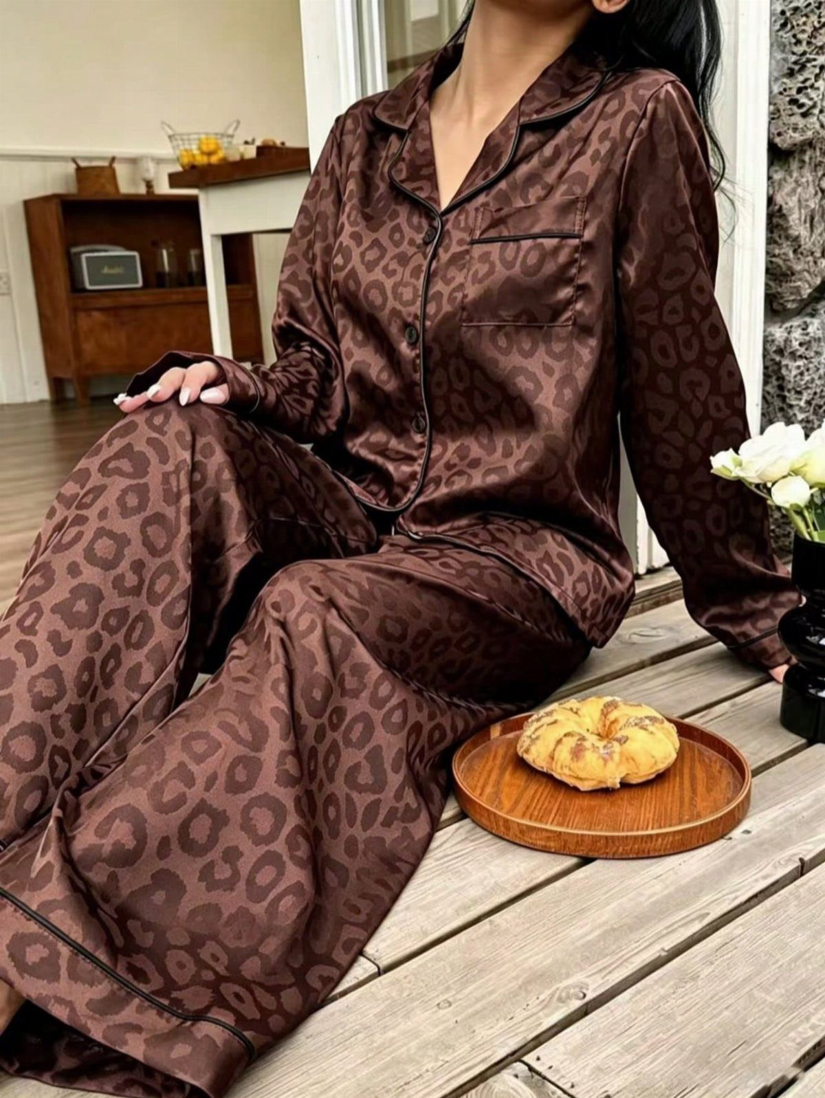 Person wearing brown leopard-print satin pajamas sitting on a wooden deck with pastry.