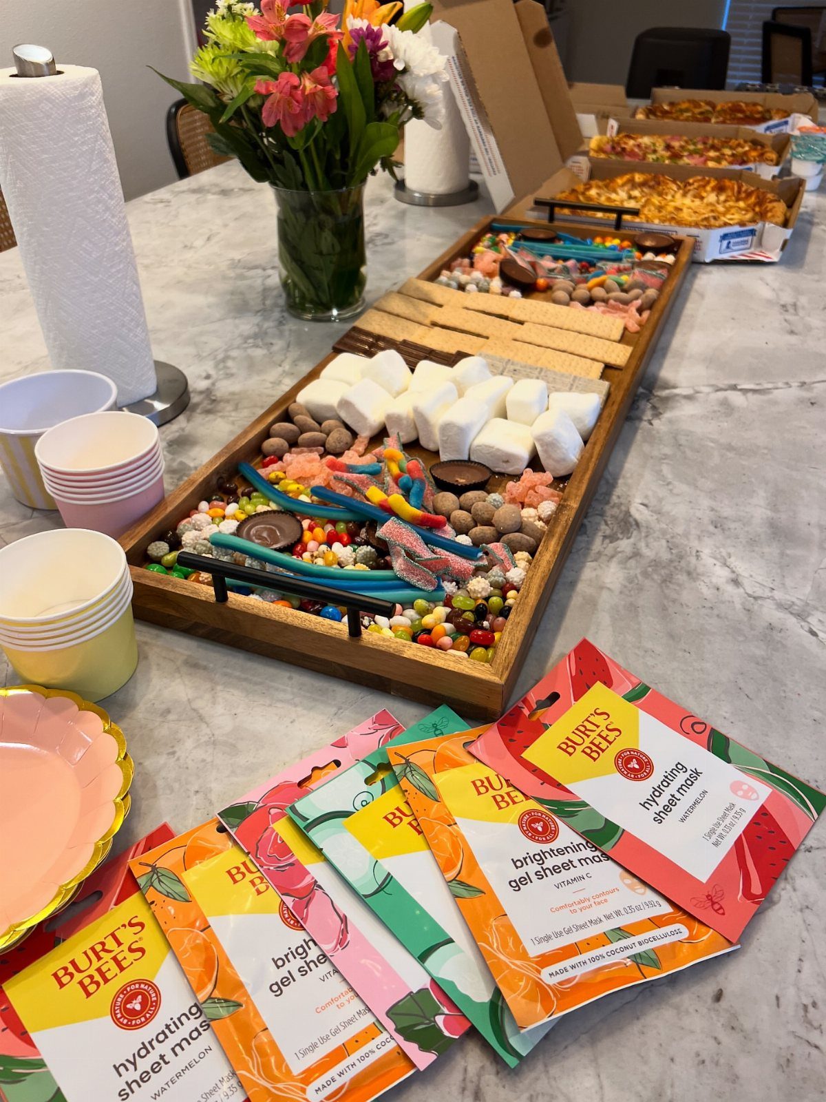 Wooden candy tray with marshmallows and chocolates next to colorful face mask packets.
