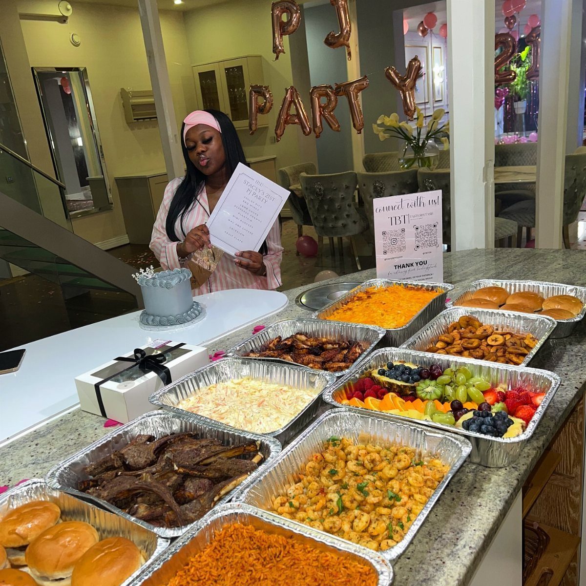 Woman in pink pajamas standing behind a buffet of party food and gold balloons.
