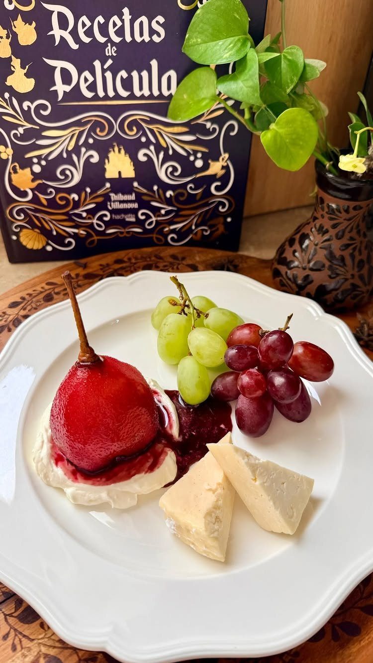 Poached red pear with grapes and cheese on a white plate next to a cookbook.