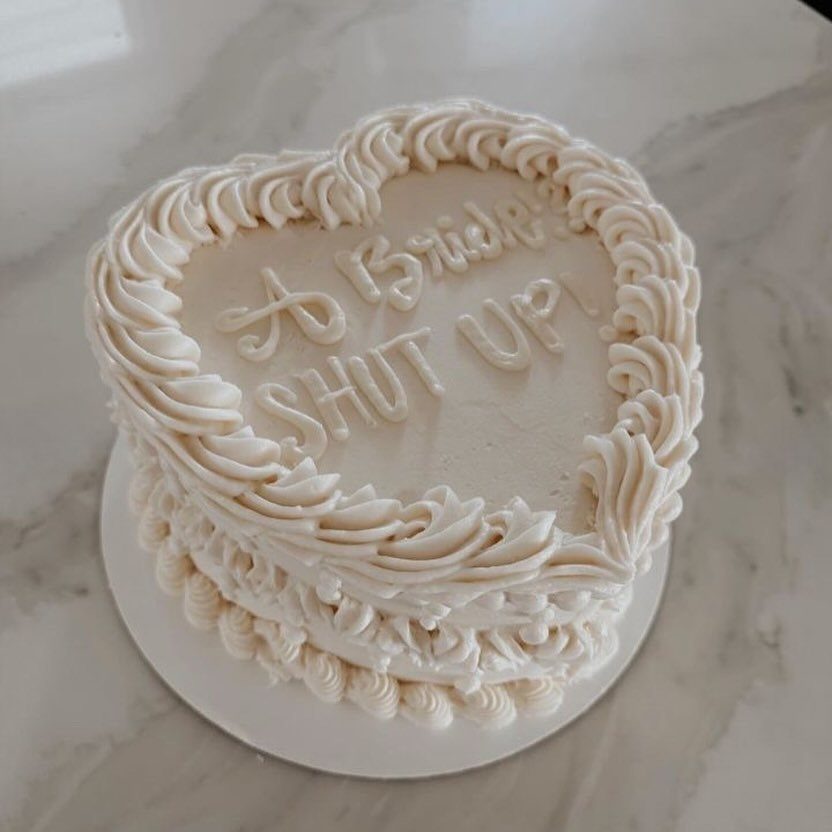 Heart-shaped white cake with piped frosting and bridal themed text on marble surface