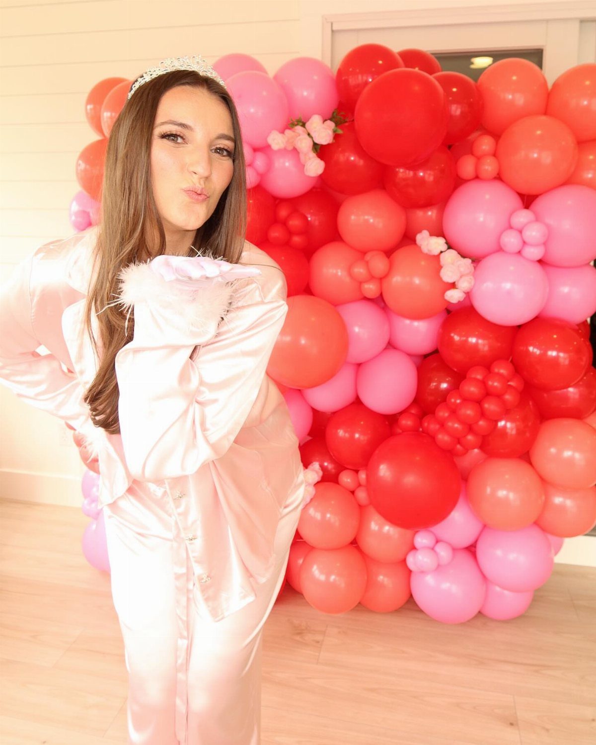 Woman in pink satin pajamas and tiara posing before pink and red balloons.