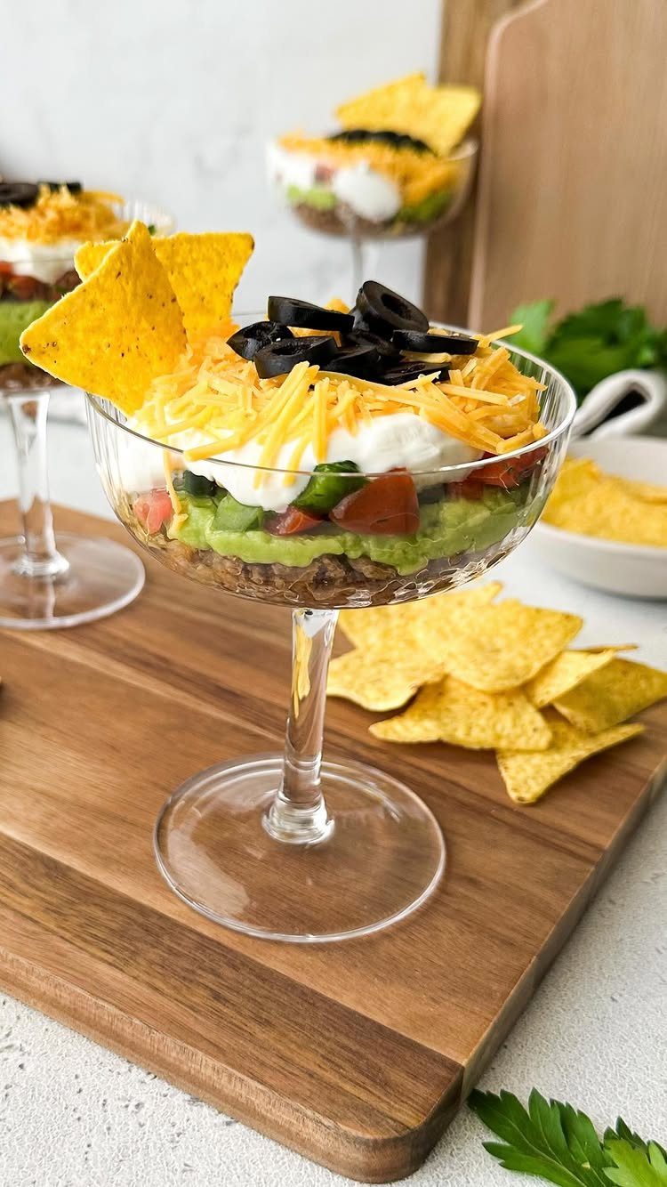 Layered taco dip in a glass coupe with tortilla chips on wooden board