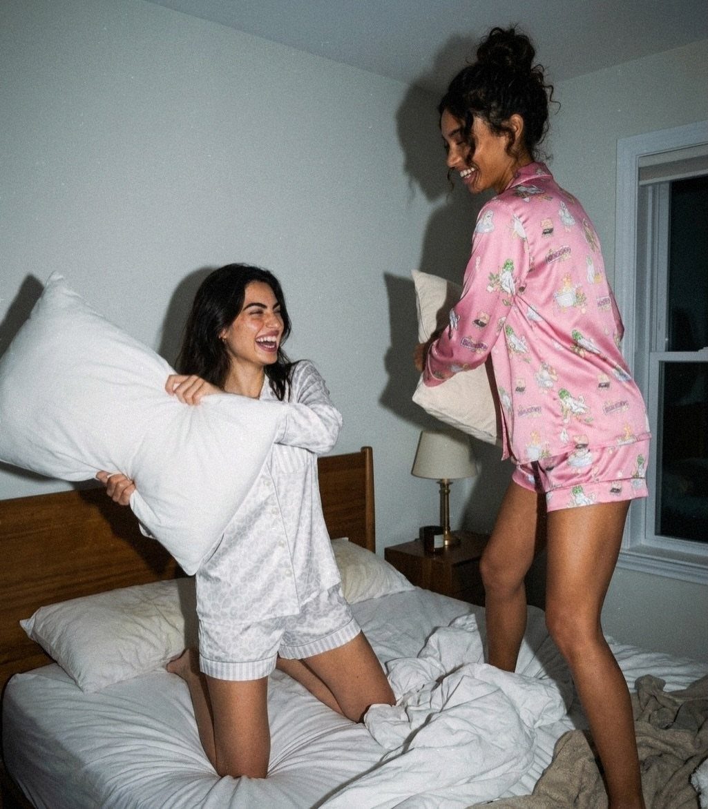 Two women in pajamas laughing and holding pillows during a pillow fight on a bed.