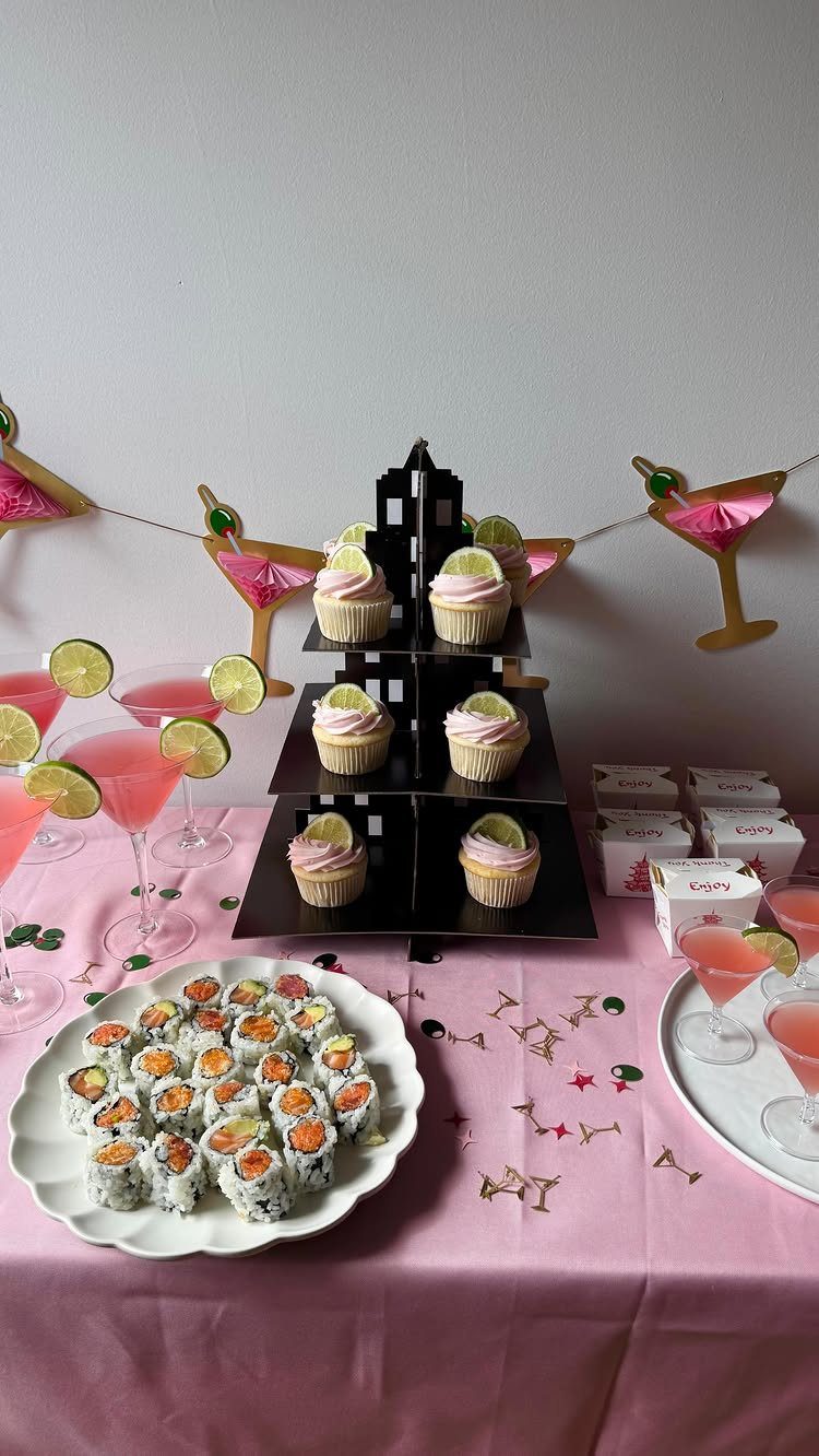 Party table with pink cocktails, sushi, and cupcakes on a pink tablecloth.