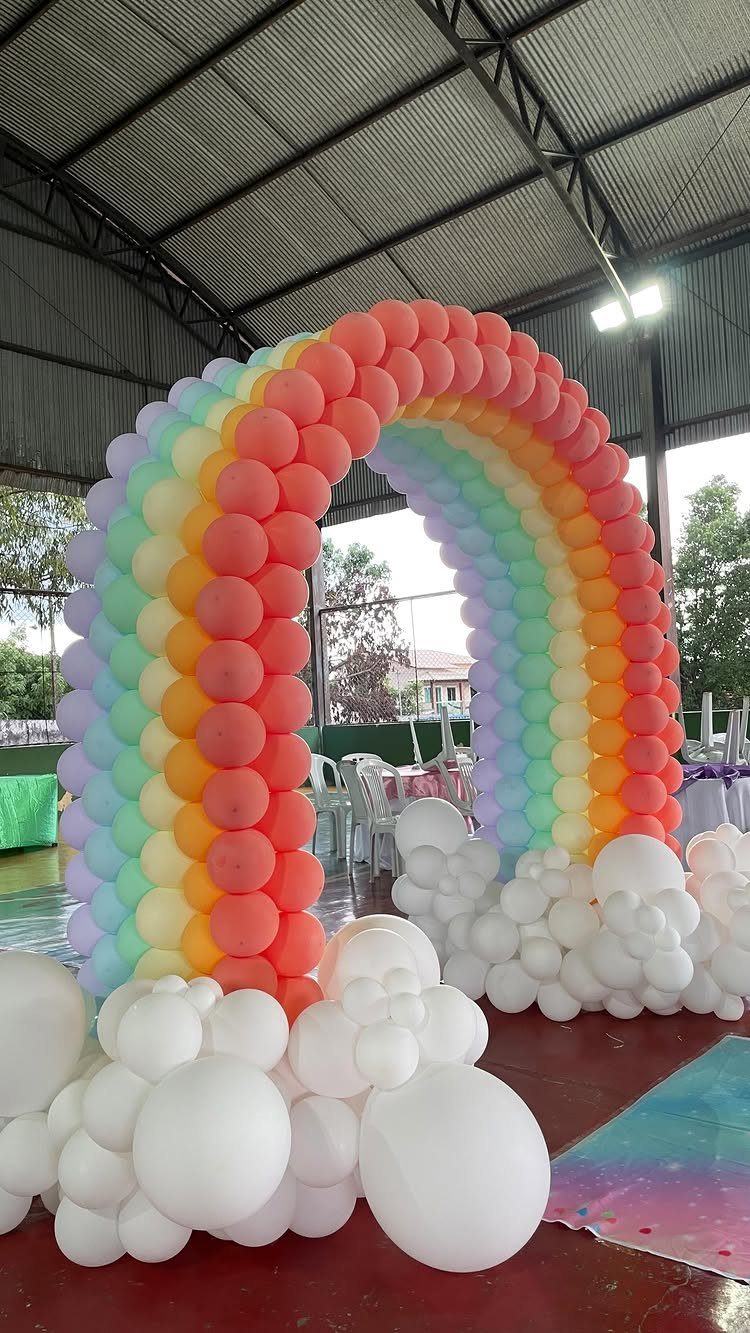 Rainbow balloon arch with white balloon clouds at the base under a pavilion.