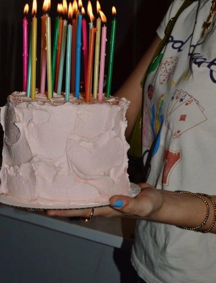 Pink frosted birthday cake with many lit rainbow colored candles held by a person