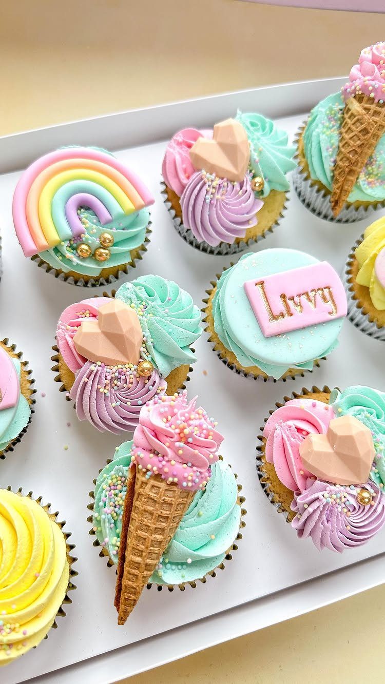Pastel rainbow cupcakes with frosting, mini cones, and hearts on a white tray.