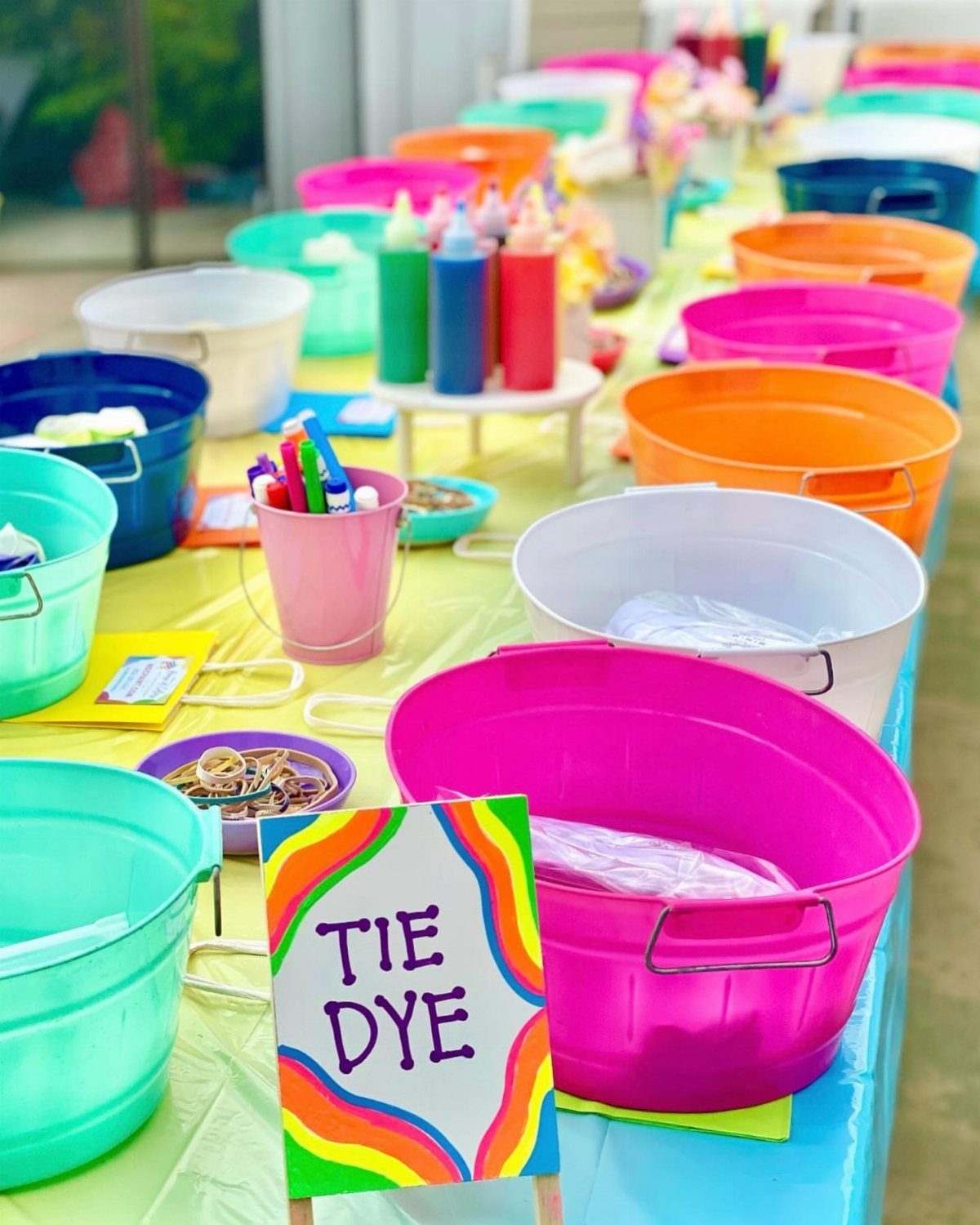 Tie dye station with colorful plastic tubs and a sign on a yellow table.