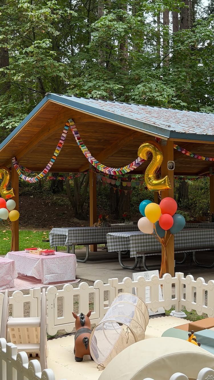 Outdoor birthday party setup under a pavilion with balloons and a white picket fence.