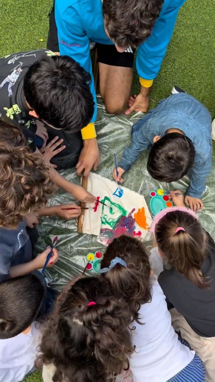 Children and an adult painting a large colorful canvas on a green lawn.