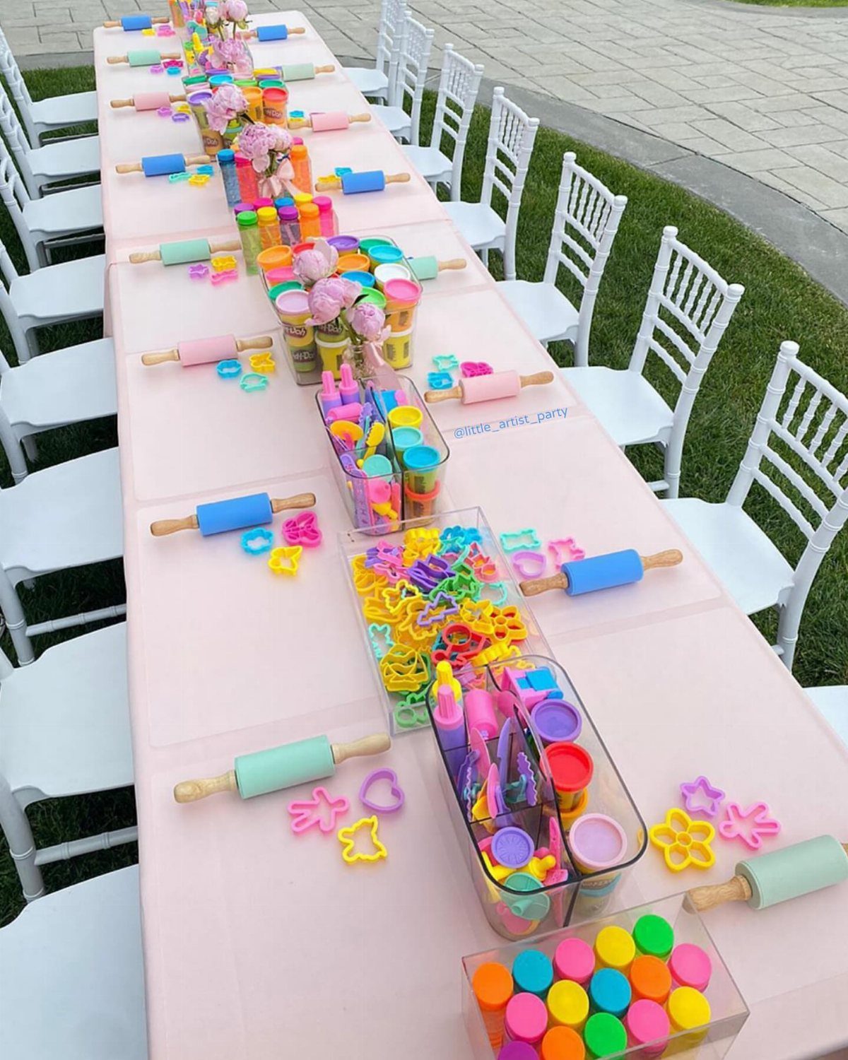 Long pink table set with colorful play-dough, rolling pins, and cookie cutters outdoors.