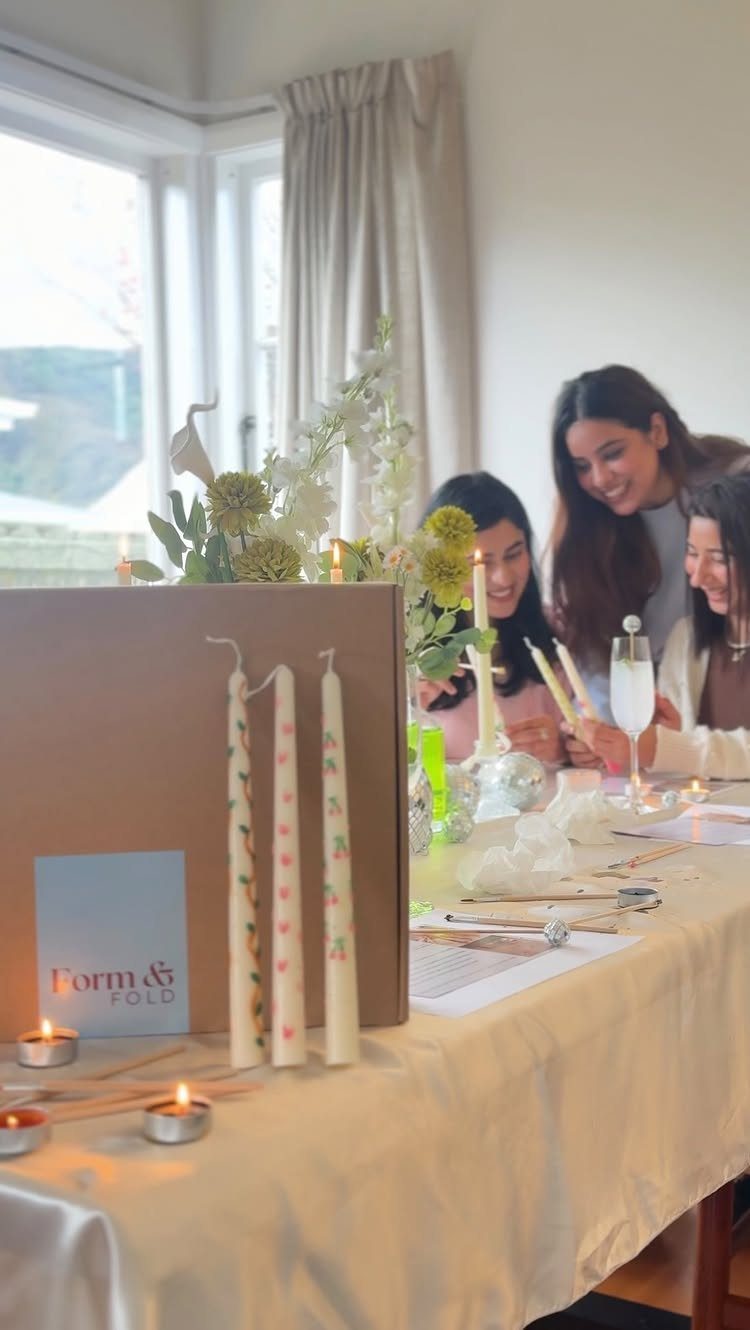 Three women decorating white taper candles at a table with floral arrangements.