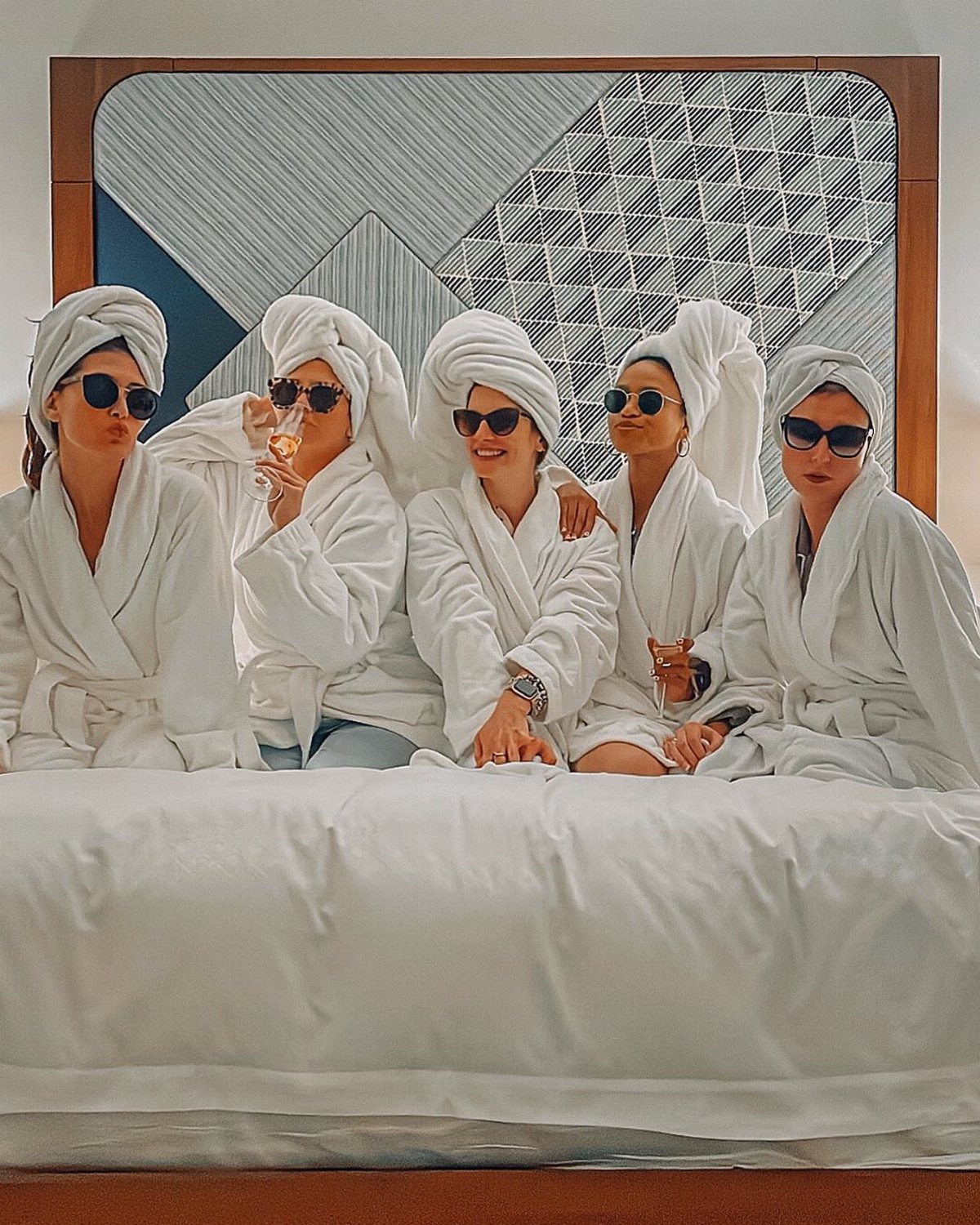 Five women in white robes and towel wraps posing on a white bed.