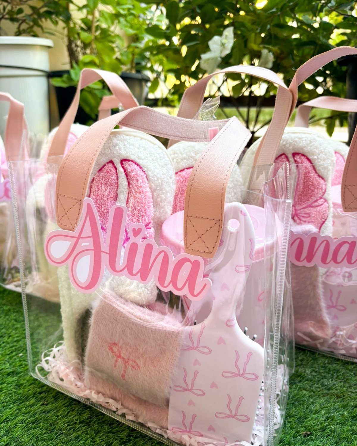 White fluffy slippers with pink accents inside clear personalized gift bags on grass.