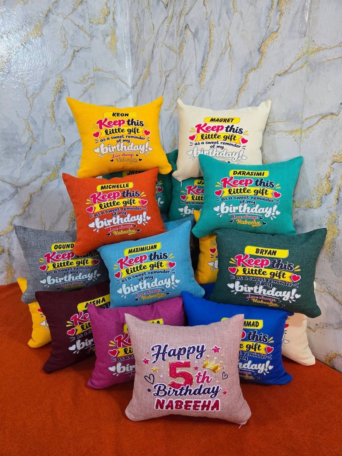 Colorful personalized birthday pillows with names and messages piled on an orange surface.