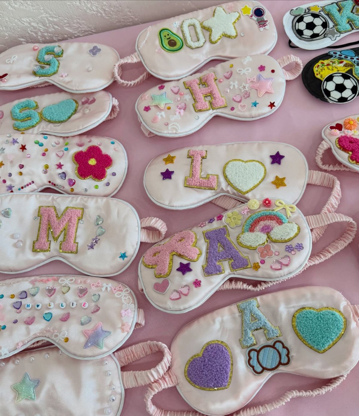 Pink satin sleep masks decorated with colorful glittery letters and whimsical shapes.