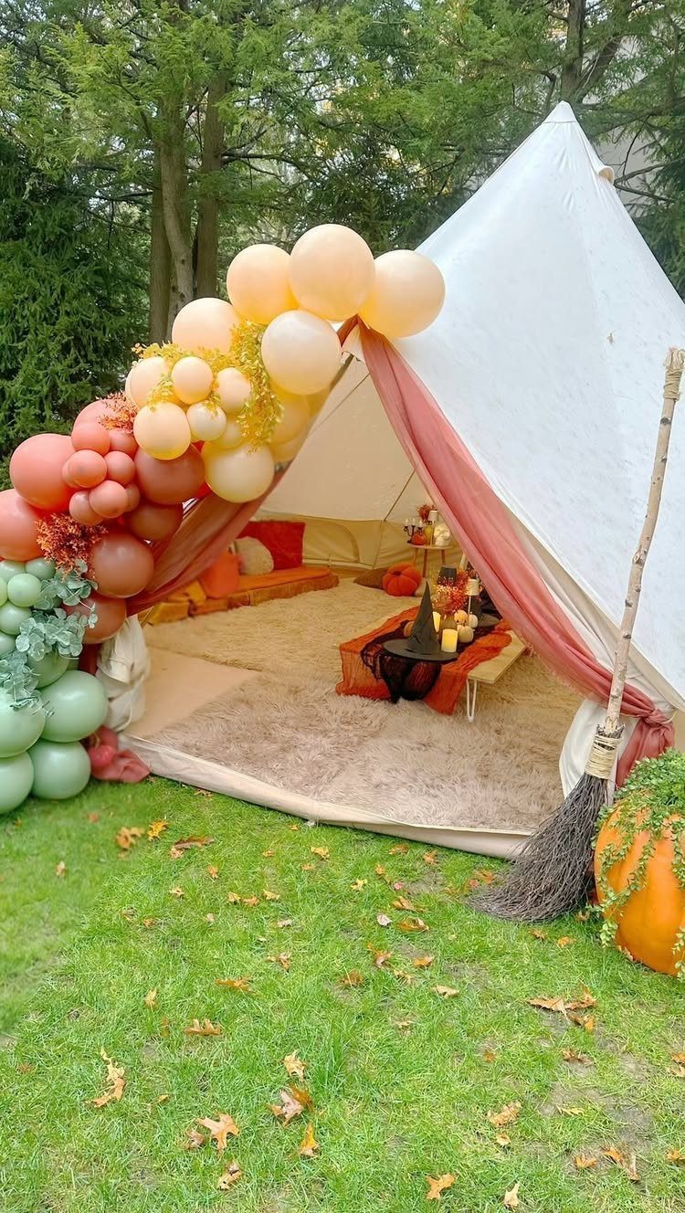 White teepee tent with autumn balloons, a broom, and pumpkin on green grass.
