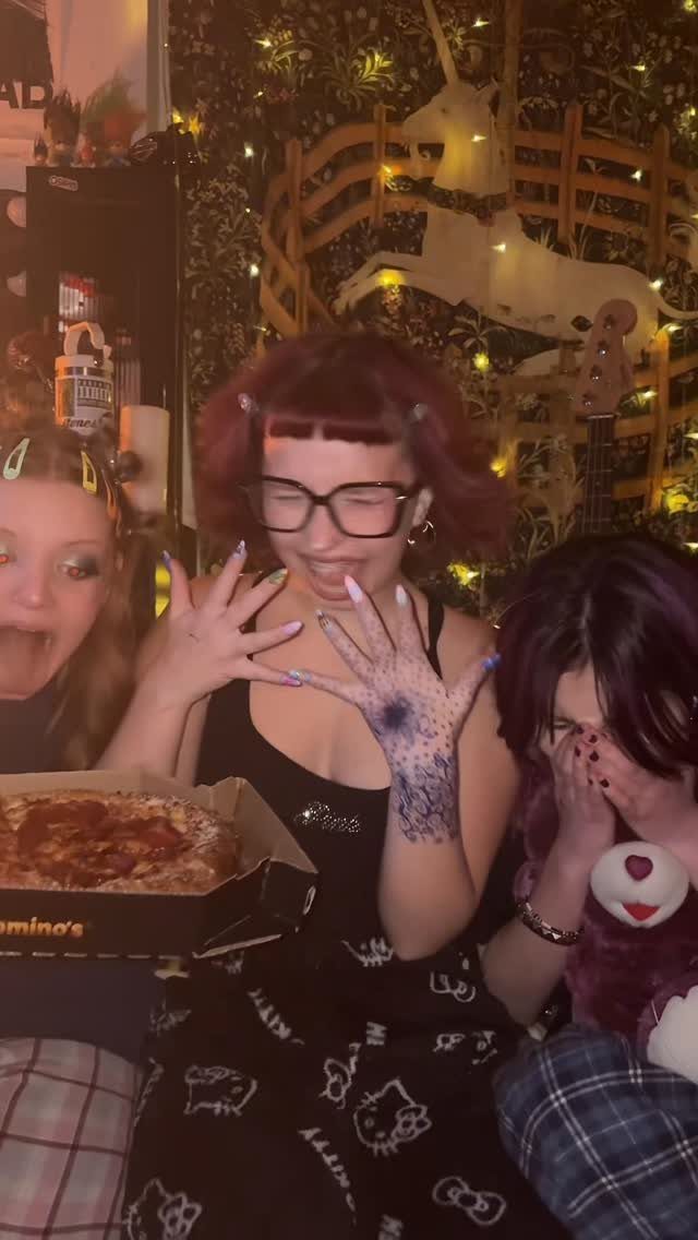 Three young women in pajamas eating pizza in a room with fairy lights.