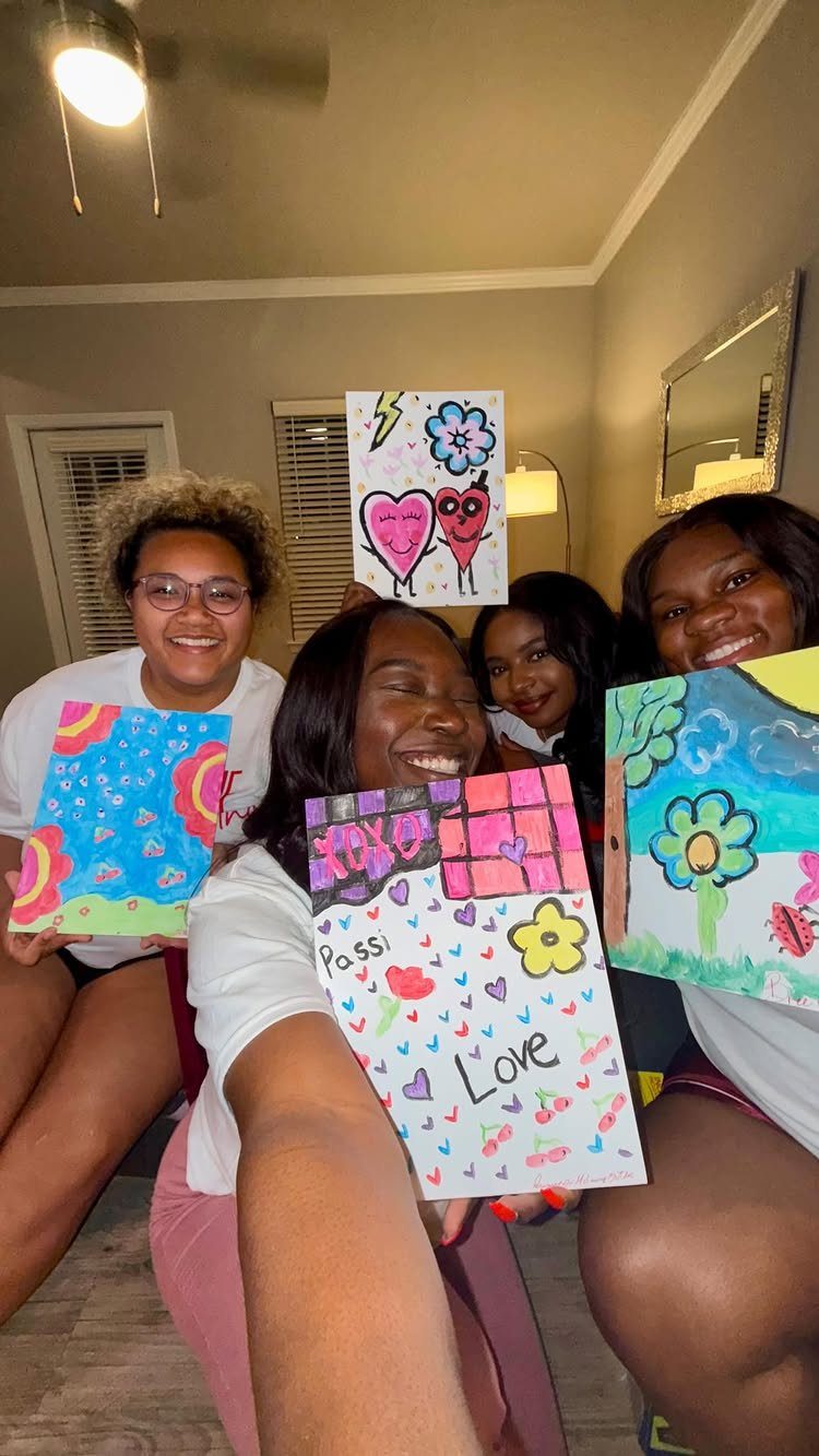 Four young women smiling and holding up their colorful hand-drawn paintings during a party.