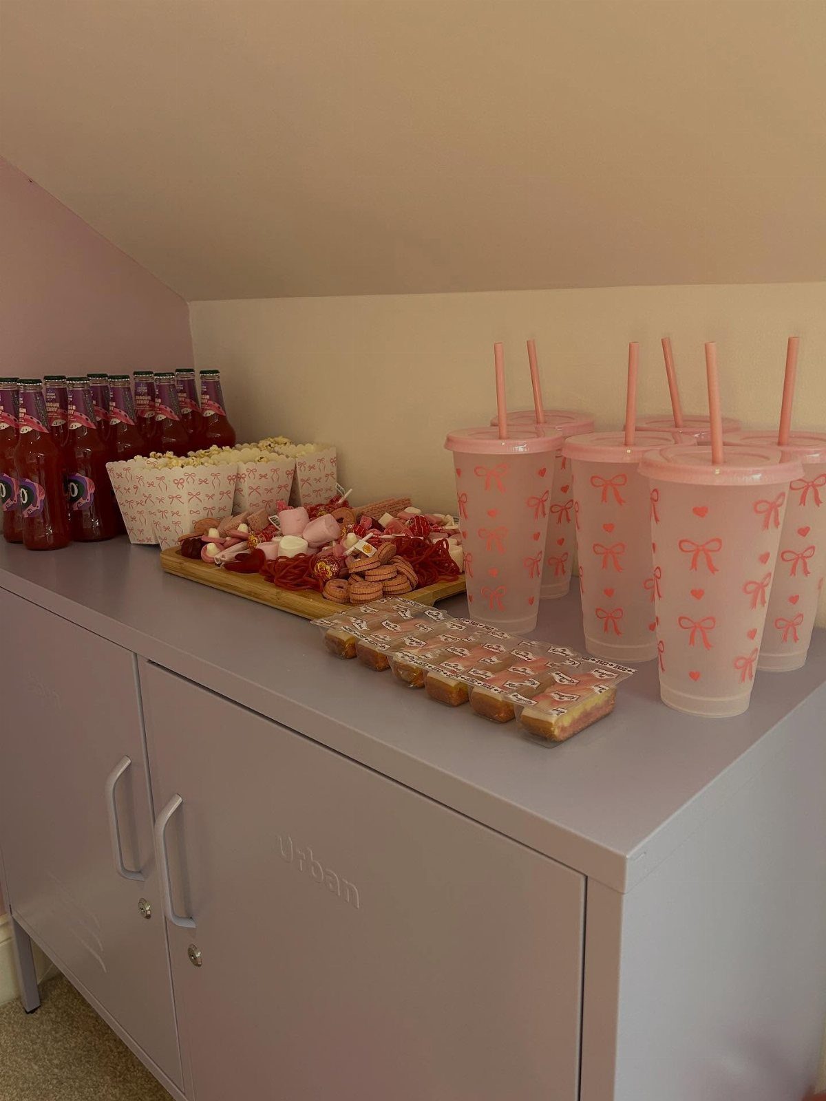 Party snacks and pink tumblers arranged on a grey cabinet for a celebration.