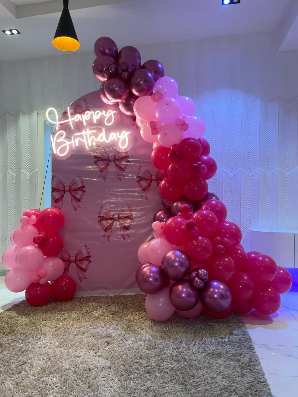 Pink birthday backdrop with neon sign, purple and pink balloons, and decorative bows.