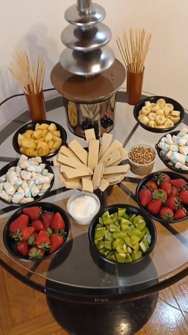 Chocolate fountain surrounded by bowls of fresh fruit, marshmallows, and wafers on a table.