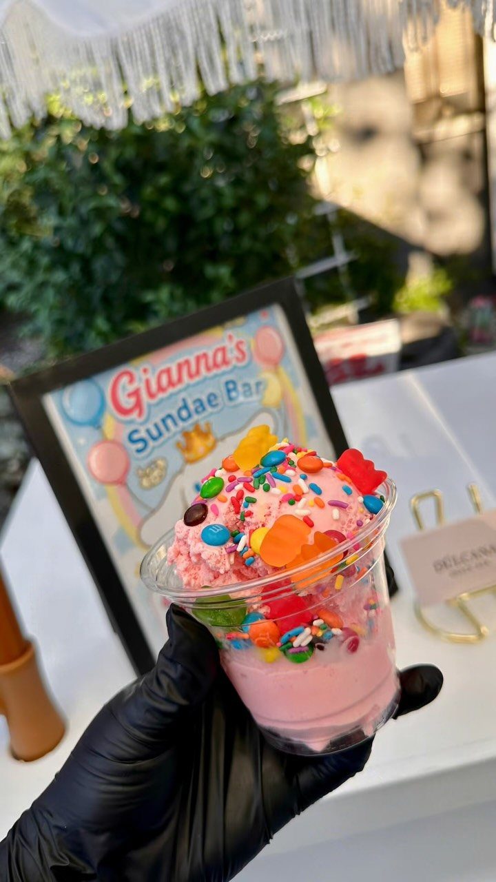 Pink ice cream sundae with colorful toppings held in front of a sundae bar sign