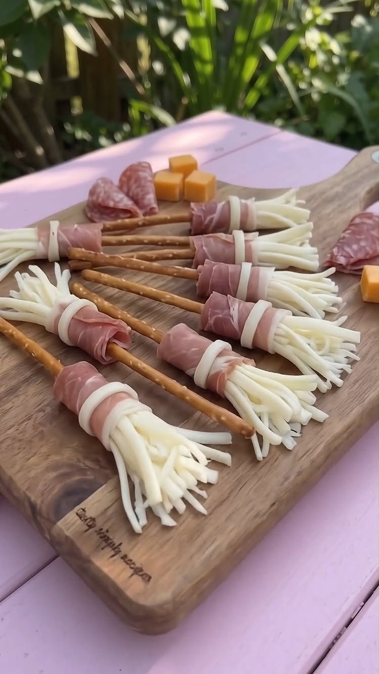 Broom-shaped snacks made of breadsticks, cured meat, and shredded cheese on wooden board.