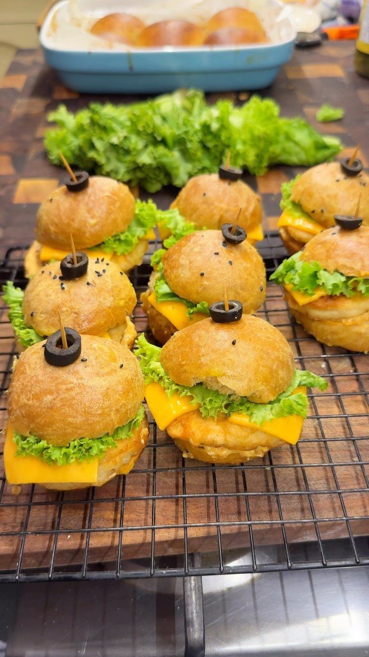 Mini sliders with cheese and lettuce topped with olives on a wire rack.