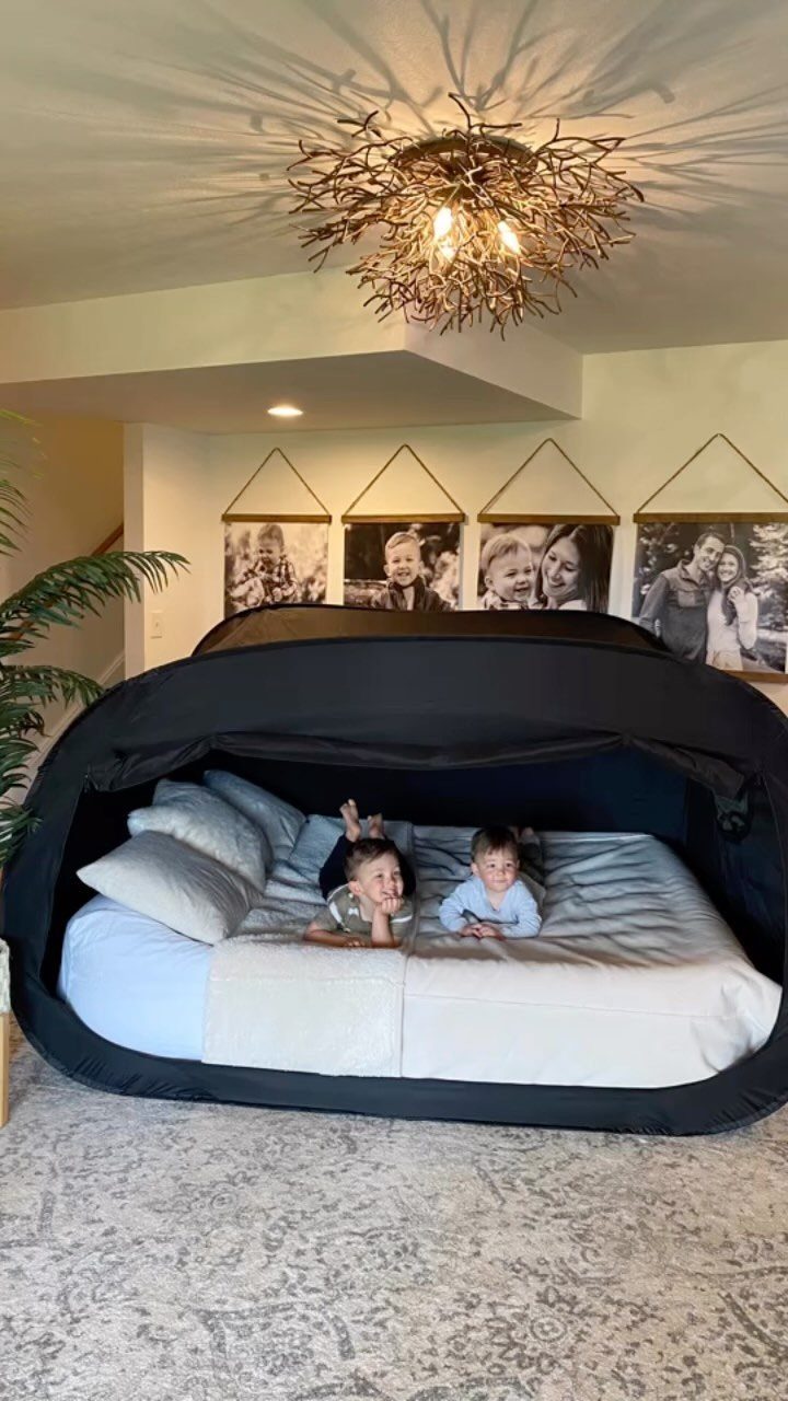 Two children lying inside a black pop-up tent over a white mattress indoors.
