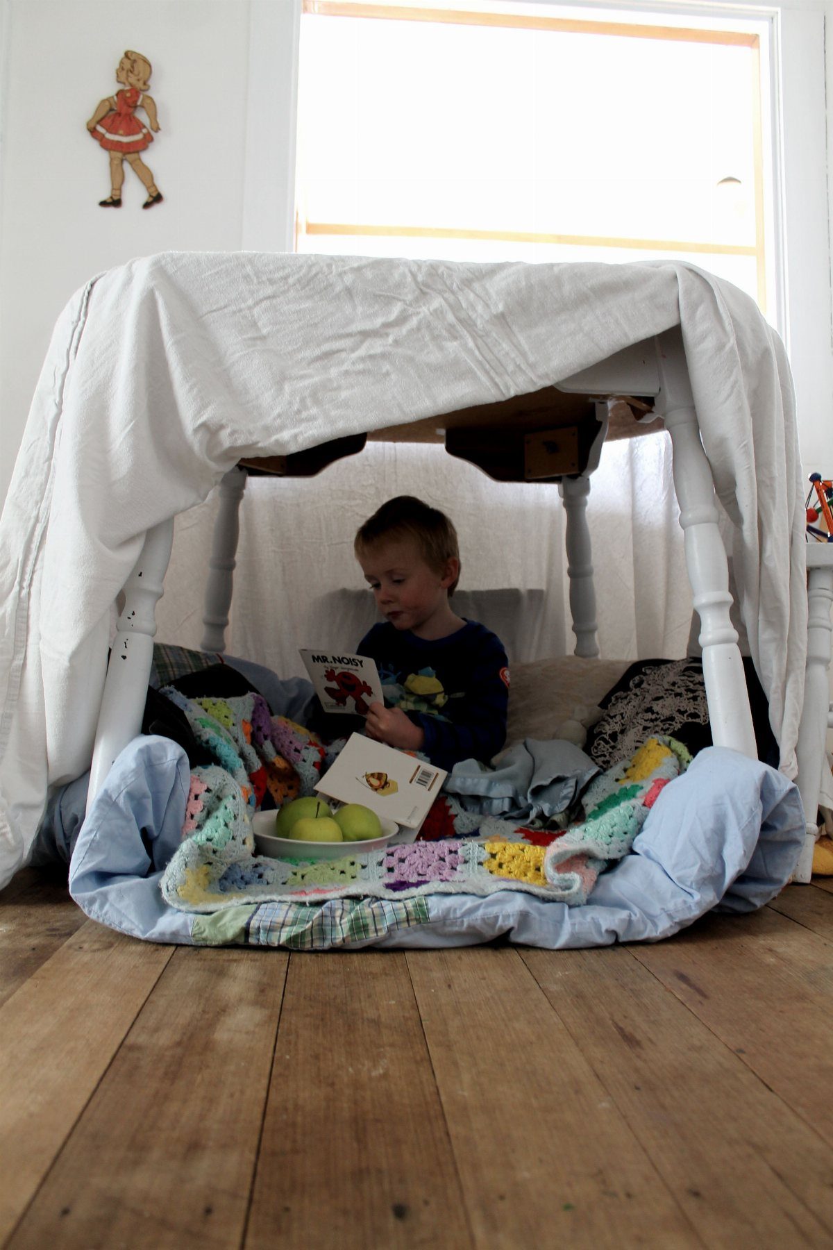 Child reading a book inside a blanket fort built under a table.