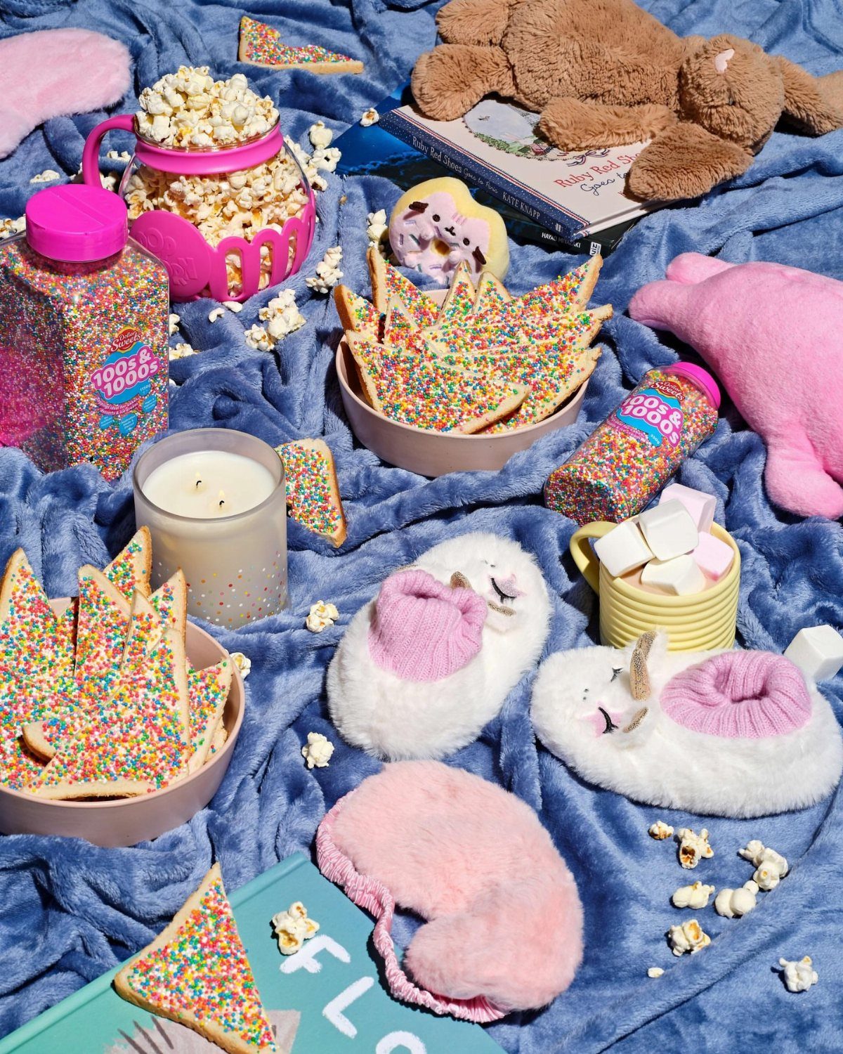 Colorful snacks, fluffy slippers, and a teddy bear on a blue plush blanket.