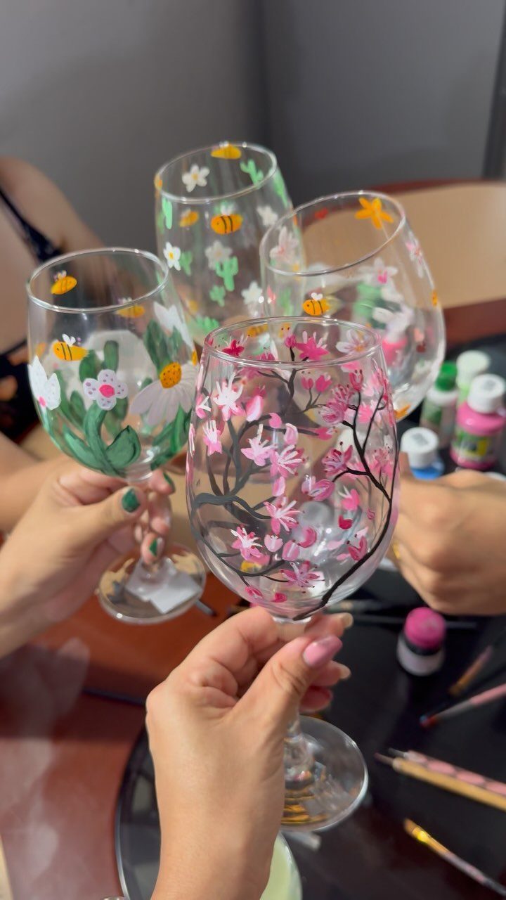 Four painted wine glasses with floral designs held up by people's hands.