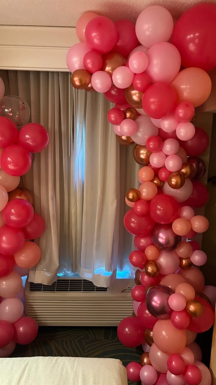 Pink and gold balloon arch framing a window with white curtains in a room.