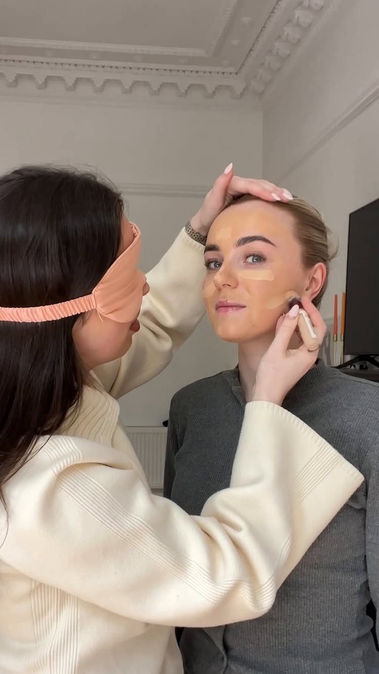 Woman wearing a pink blindfold applying makeup to another woman's face with a brush.