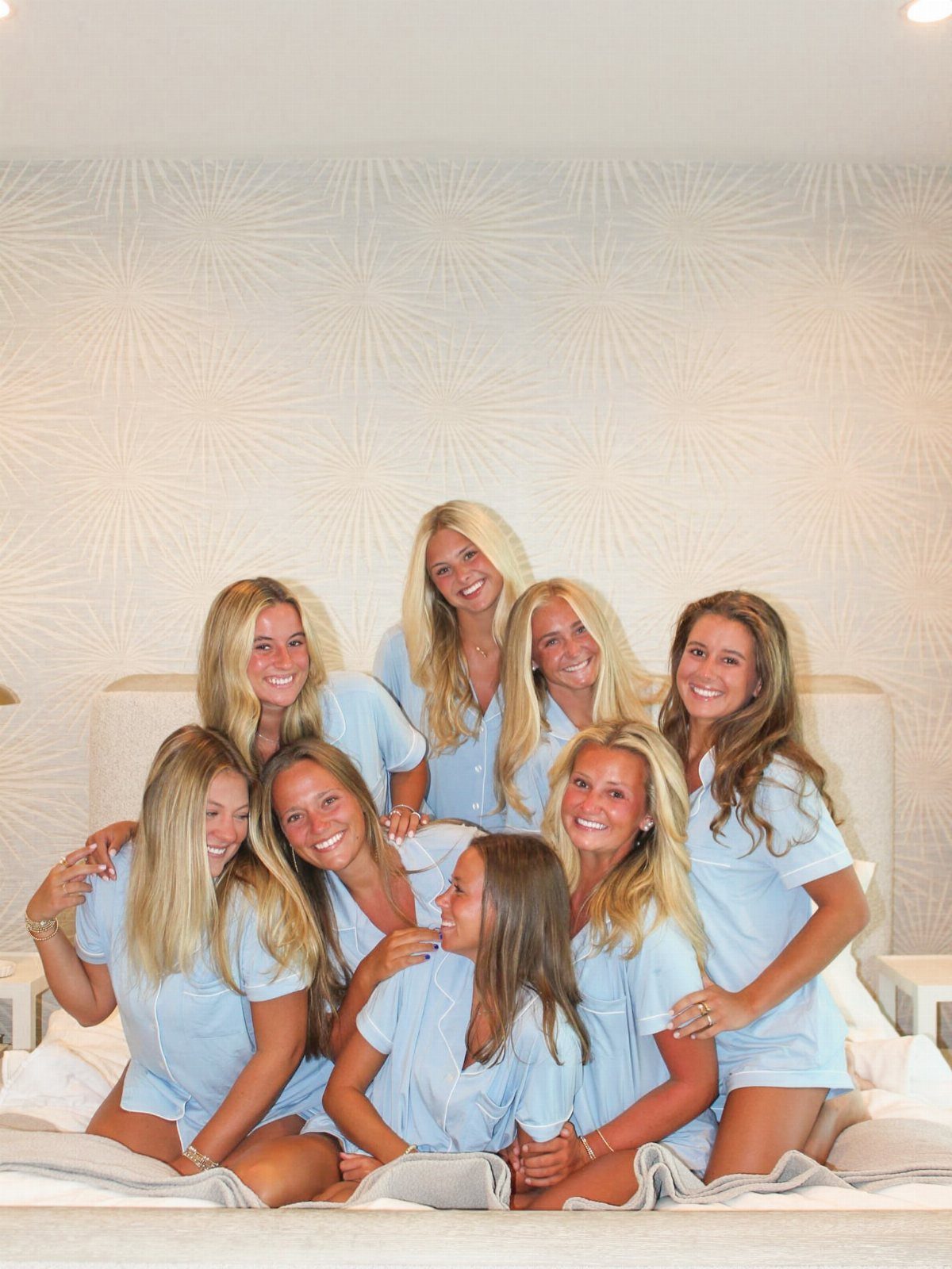Eight young women in matching light blue pajama sets posing on a white bed.
