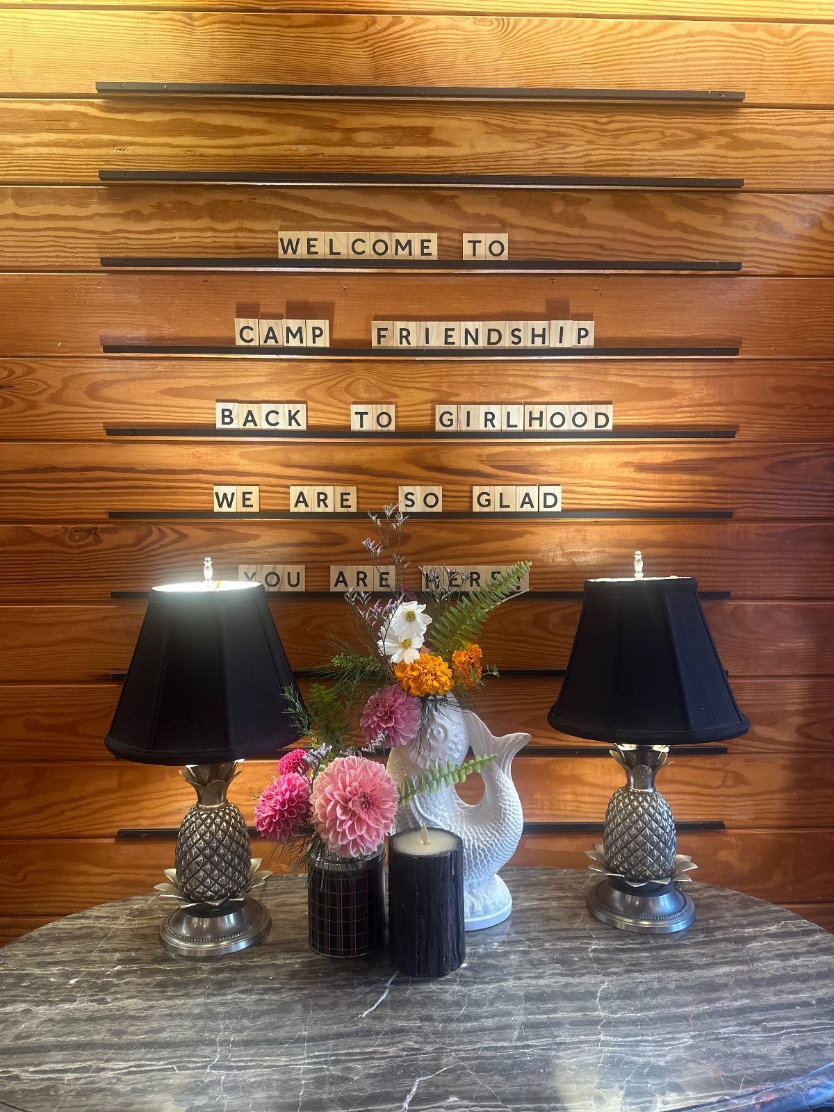 Wooden wall with welcome tiles for Camp Friendship, flanked by lamps and flowers.
