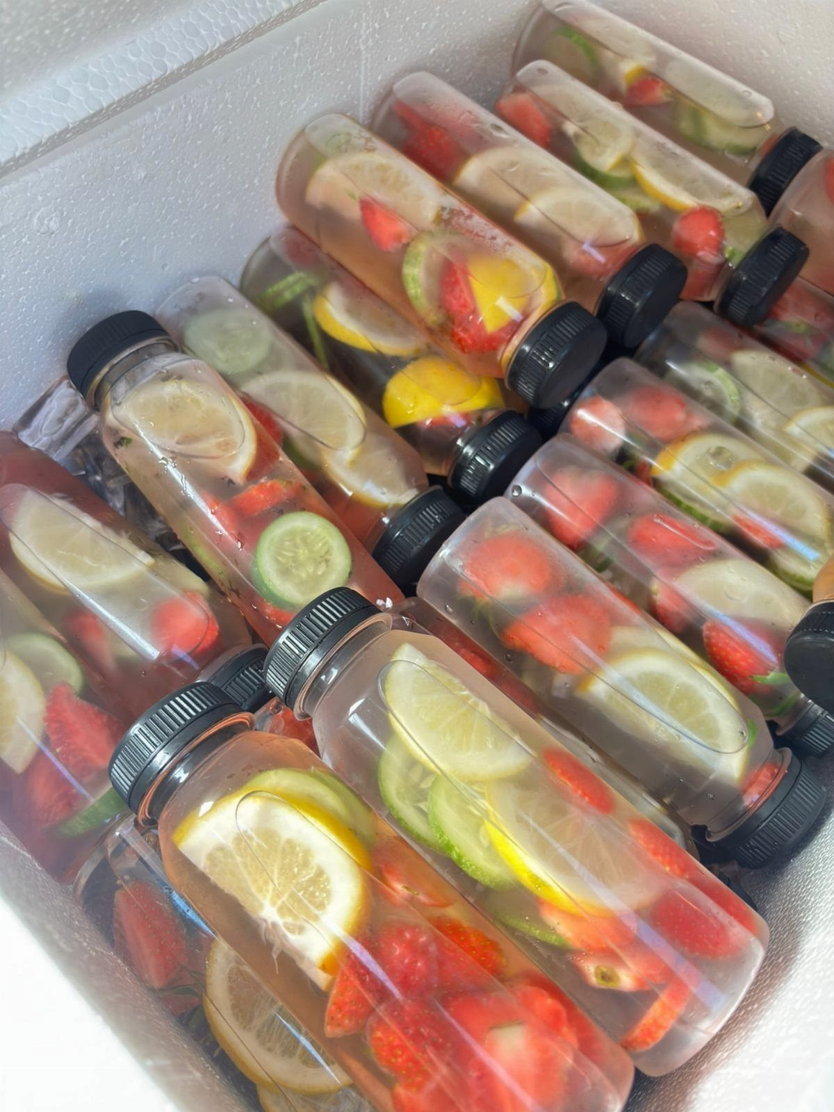 Bottles of infused water with lemon, cucumber, and strawberries in a white cooler.