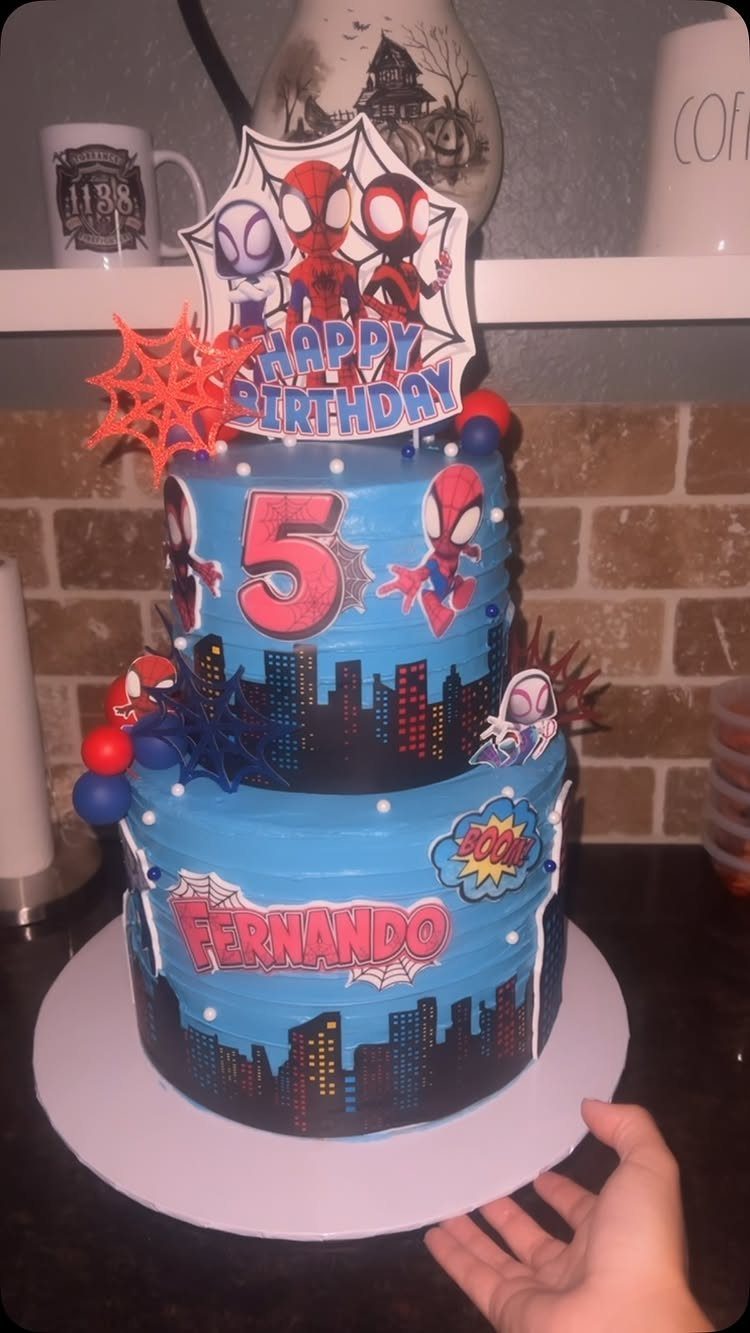 Two-tier blue Spider-Man cake with a black city skyline and birthday toppers.