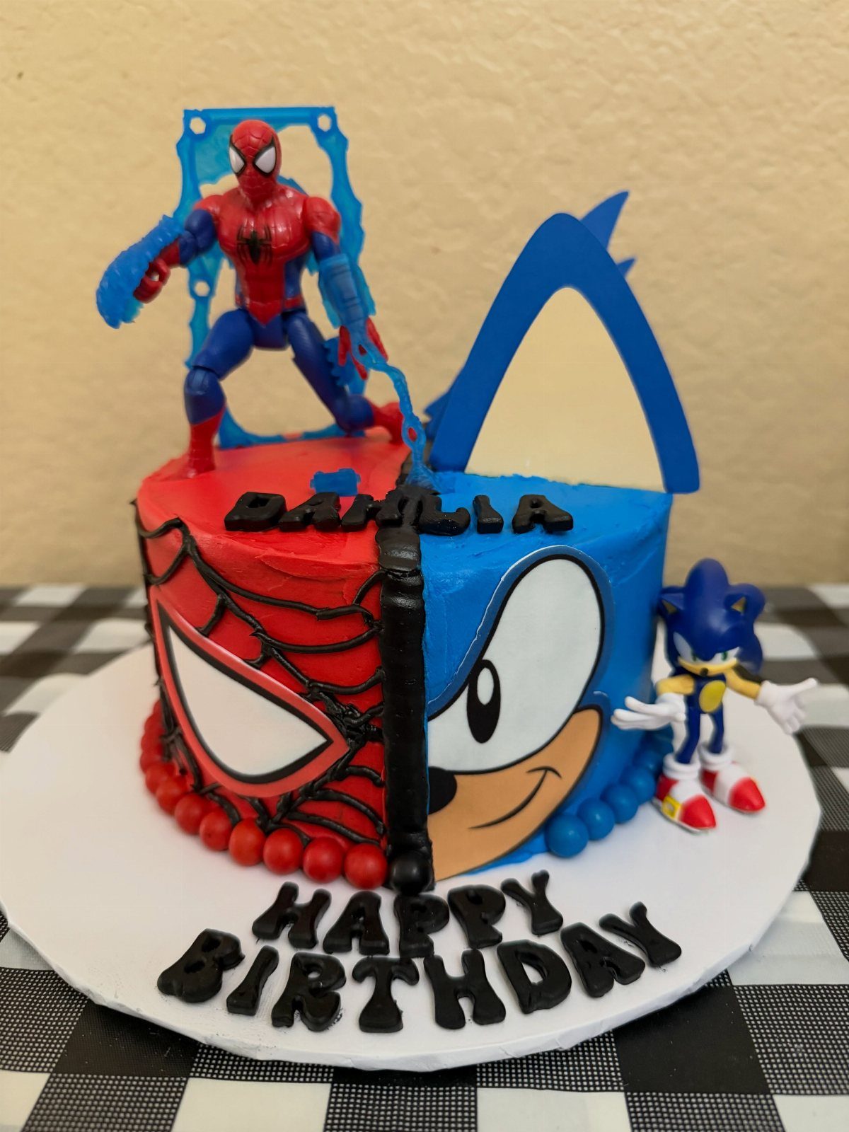 Split Spiderman and Sonic birthday cake with character figures and birthday lettering.