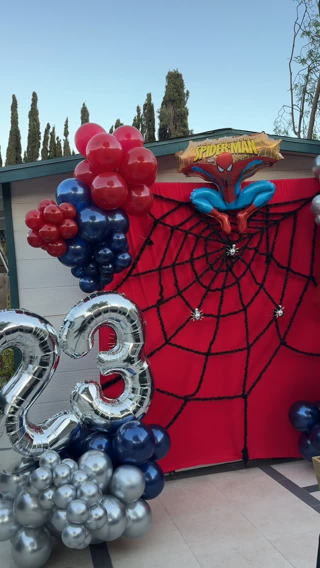 Spiderman themed photobooth backdrop with red fabric, spider web, and colorful balloons.