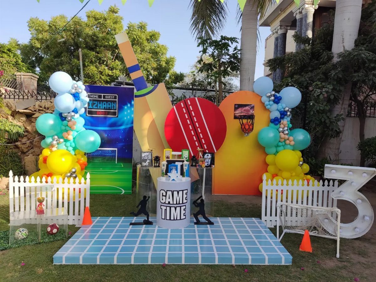 Sports themed party setup with large cricket ball, basketball hoop, and colorful balloons.