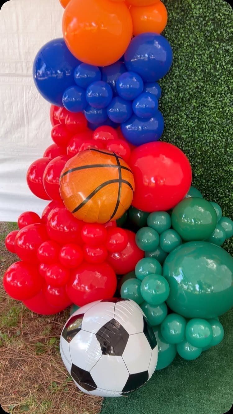 Sports-themed balloon arch with basketball and soccer ball balloons against a green hedge.