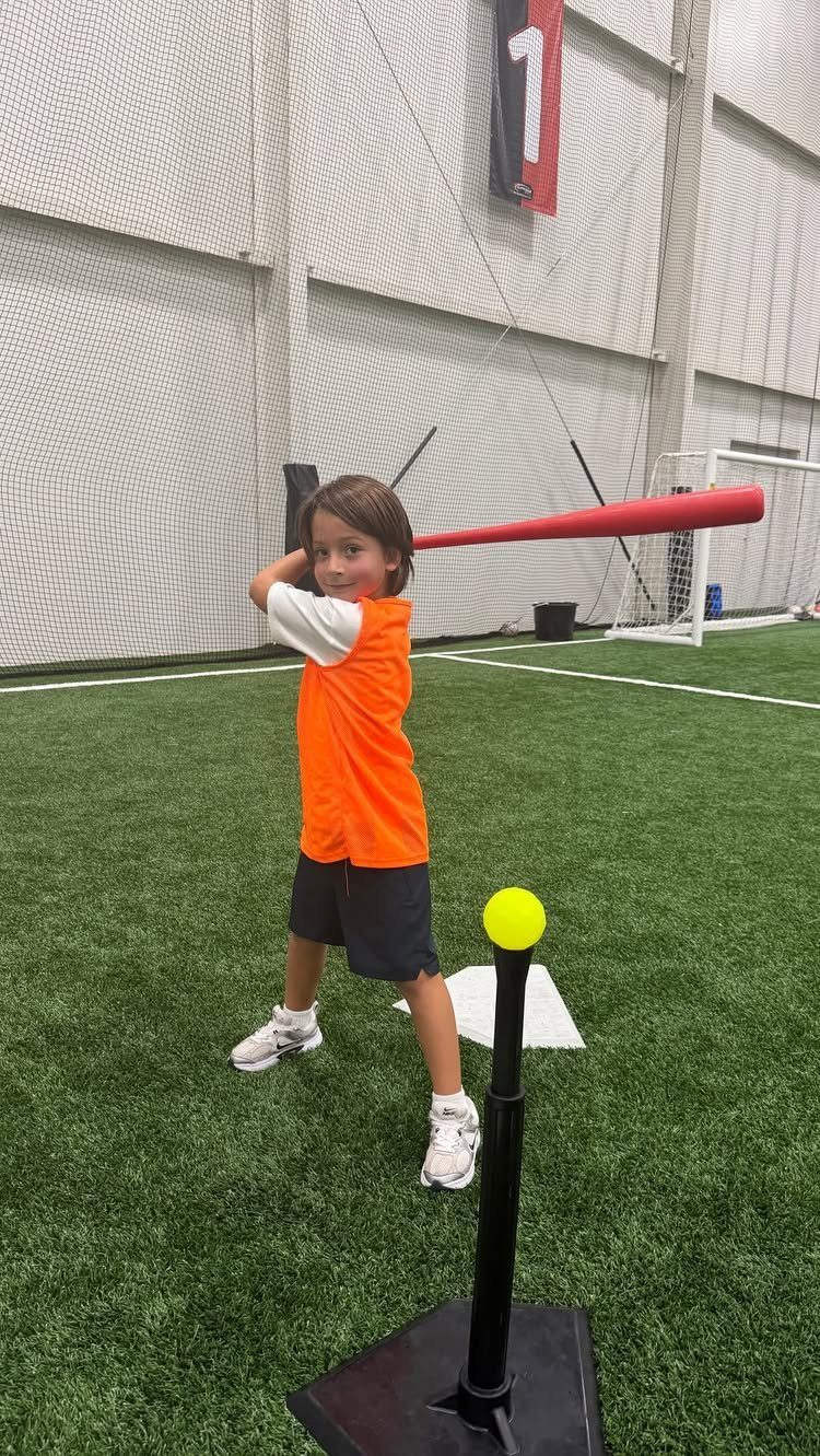 Child in orange shirt hitting a yellow ball with a red bat indoors