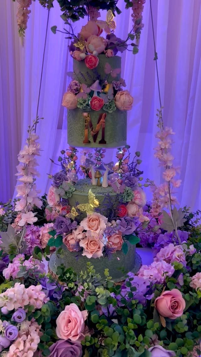 Tiered green cake with pink roses and gold butterflies surrounded by lush greenery.