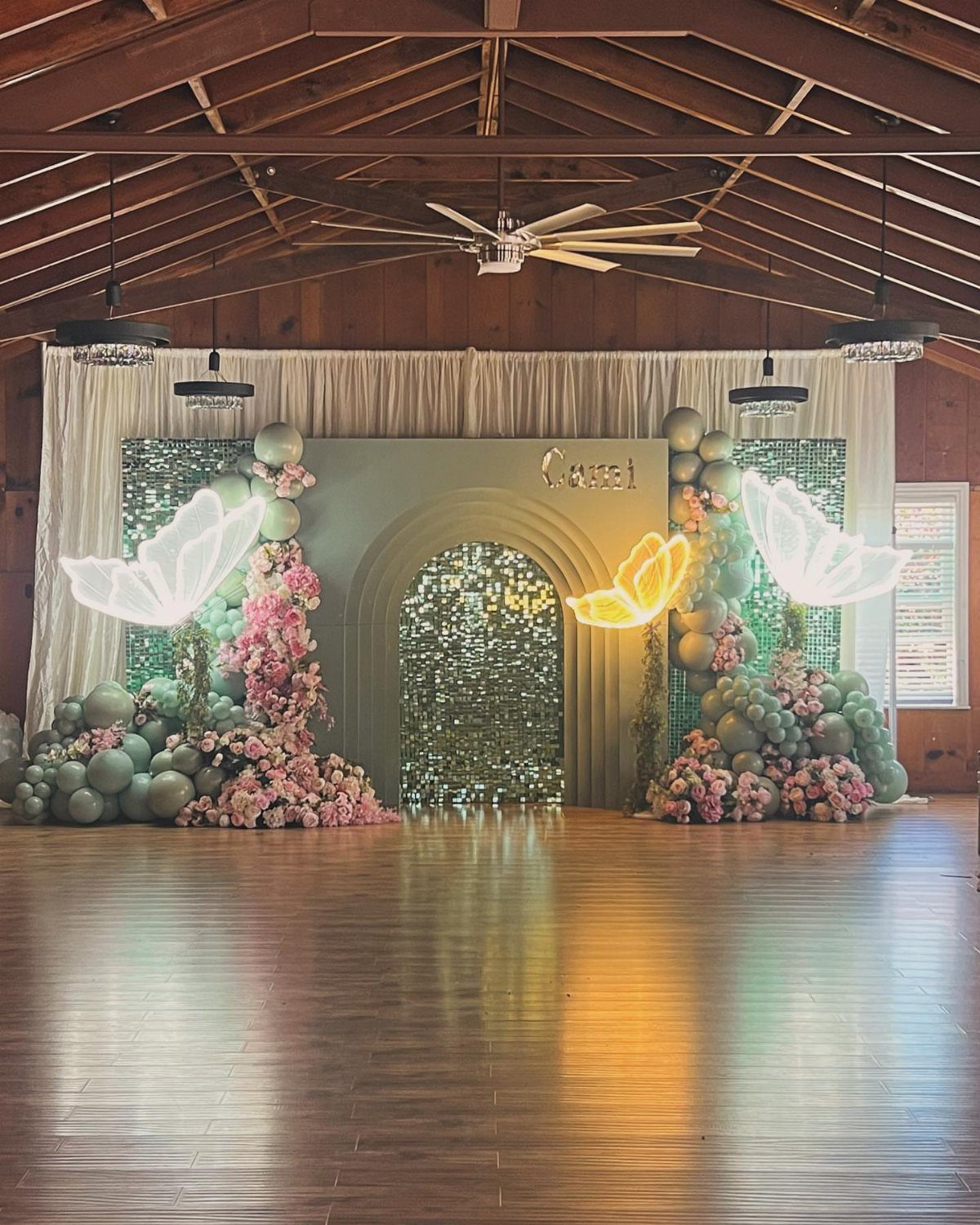 Party backdrop with green balloons, pink flowers, and glowing butterfly wing decorations.