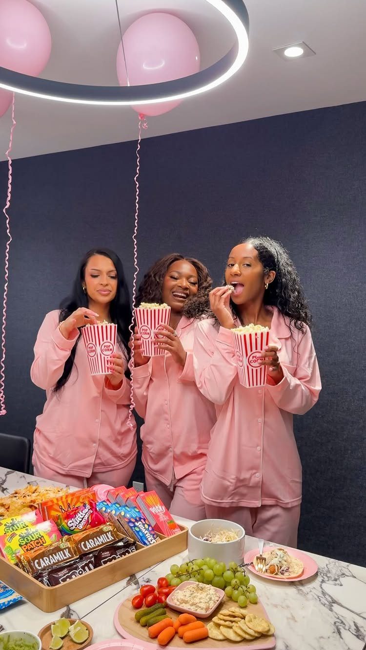 Three young women in pink pajamas eating popcorn and snacks at a party.