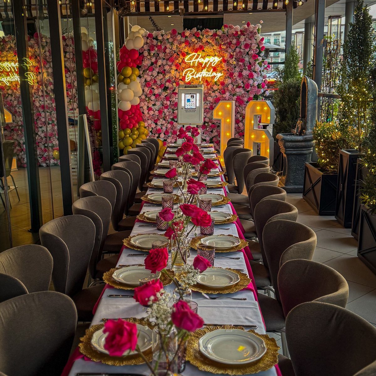 Long dinner table with pink roses and gold plates before a flower wall.